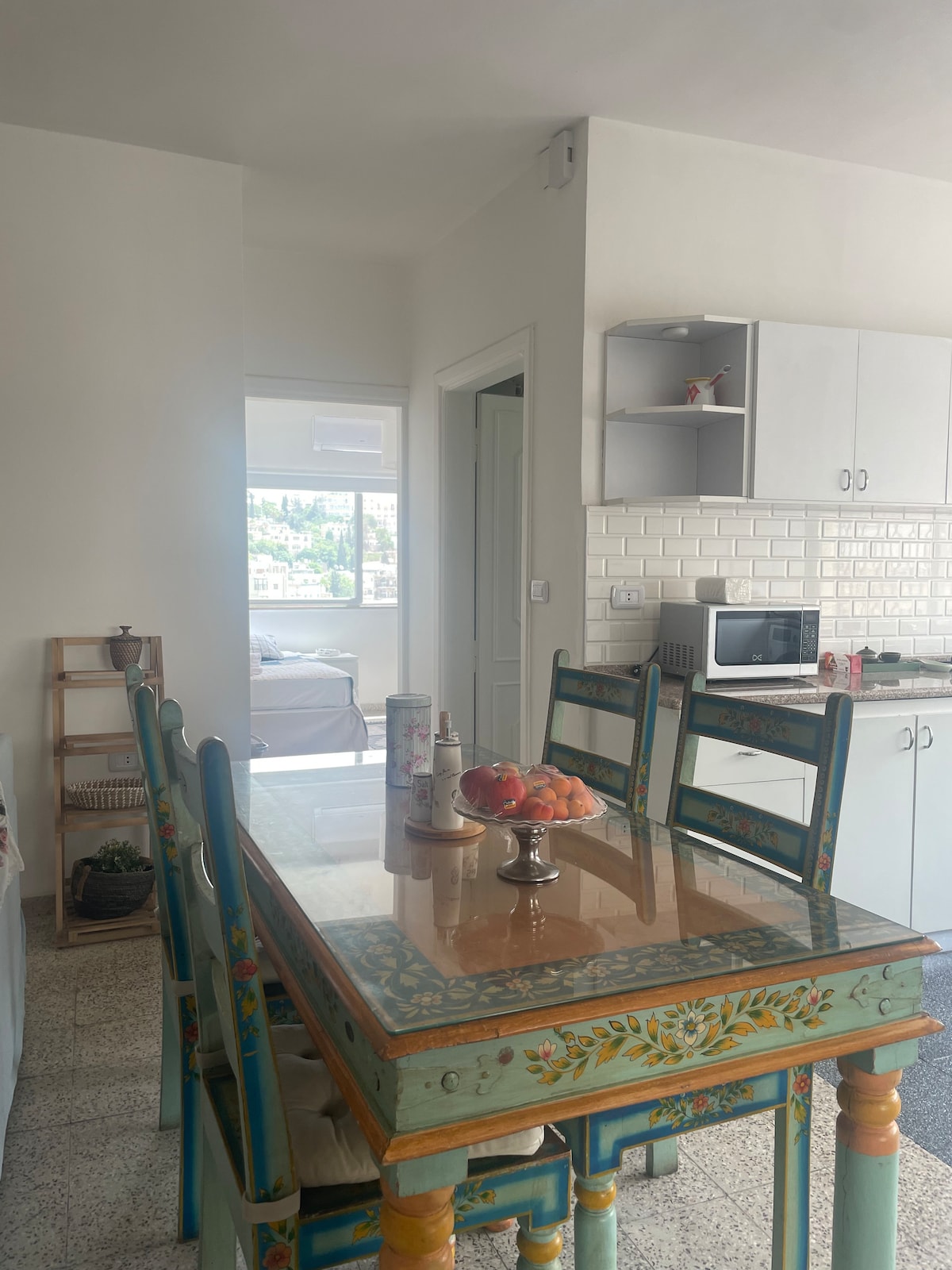 A dining area is highlighted by a wooden table featuring colorful floral designs, surrounded by matching chairs. A bowl of fresh fruit sits at the center of the table. In the background, a kitchen is visible, complete with white cabinetry and modern appliances.