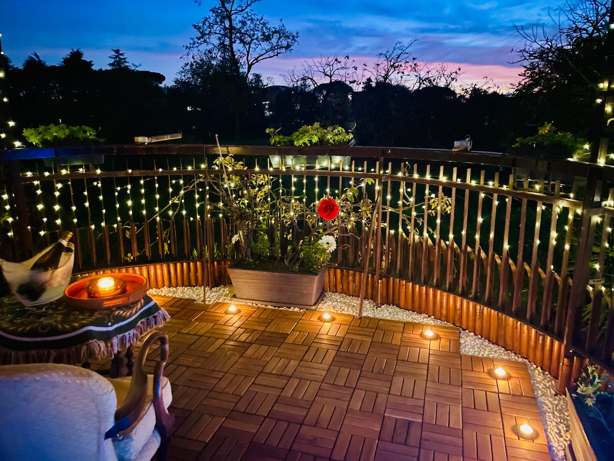 A charming terrace is illuminated by soft string lights, creating a warm ambiance against a twilight sky. Potted plants and a single red flower are arranged in a wooden planter. The wooden deck is complemented by decorative stones and cozy seating positioned nearby.