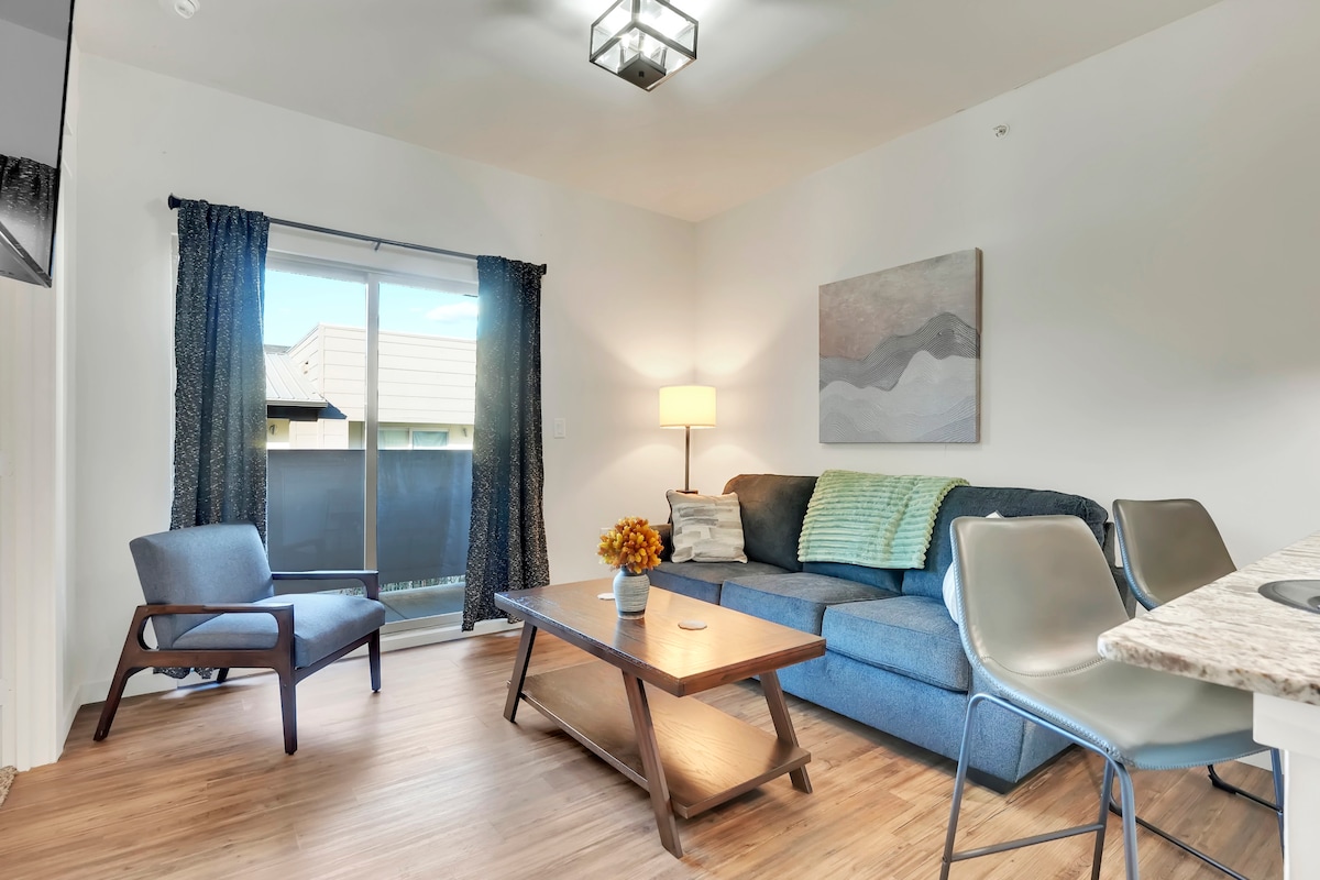 A cozy living area is presented with a blue sectional sofa and a wooden coffee table at its center. Natural light enters through large sliding glass doors, framed by decorative dark curtains. A modern lamp and a piece of wall art add character to the space.