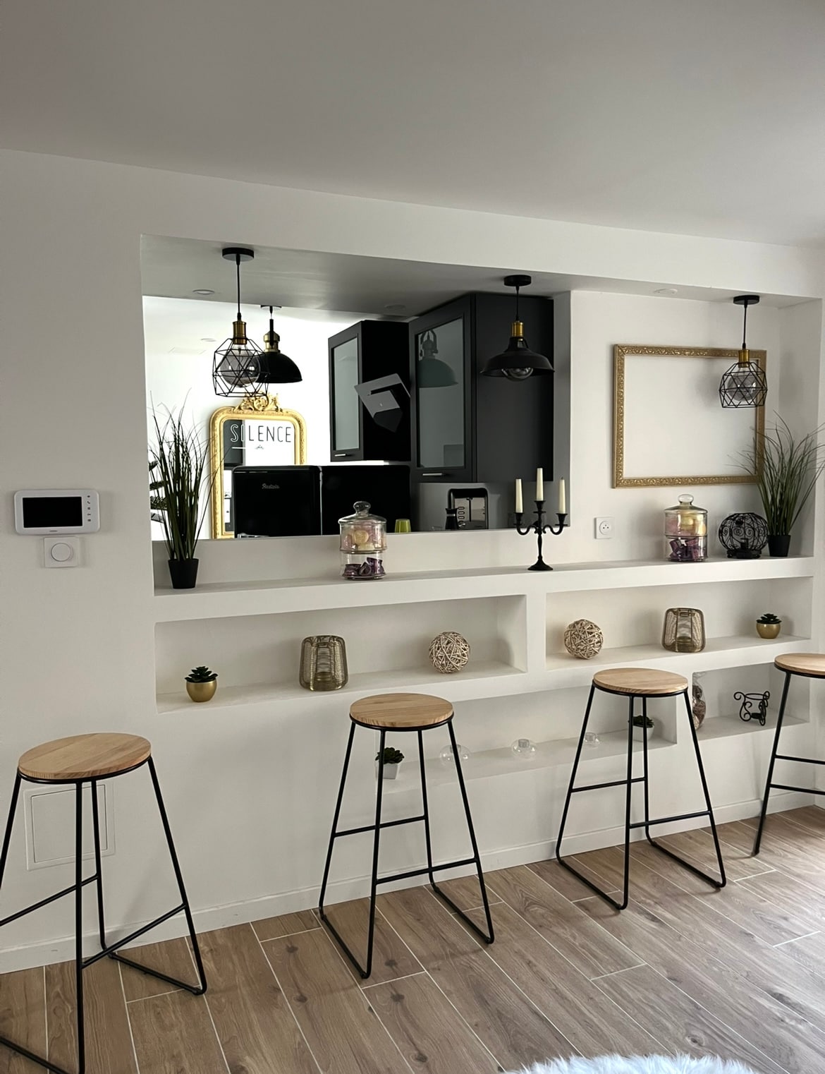 A light and airy space features a bar area with four high stools positioned at a sleek countertop. Decorative shelves display various jars and plants, while pendant lights hang above, providing soft illumination. A glimpse of the kitchen can be seen through the open design.