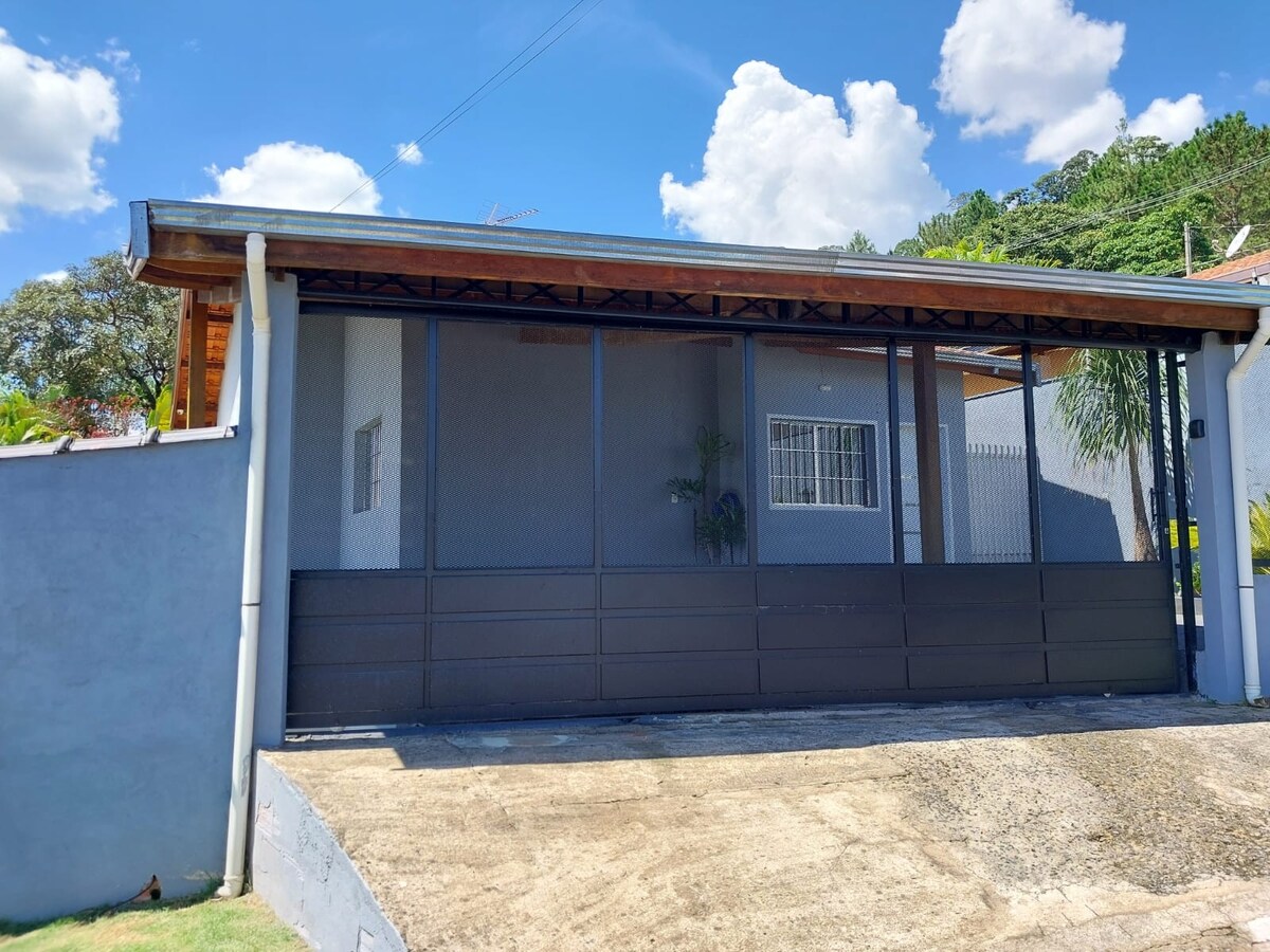 A single-story house is presented, featuring a large gate that offers a glimpse of the enclosed front yard. A window is visible, providing natural light to the home. The structure is complemented by a wooden roof and surrounded by greenery.