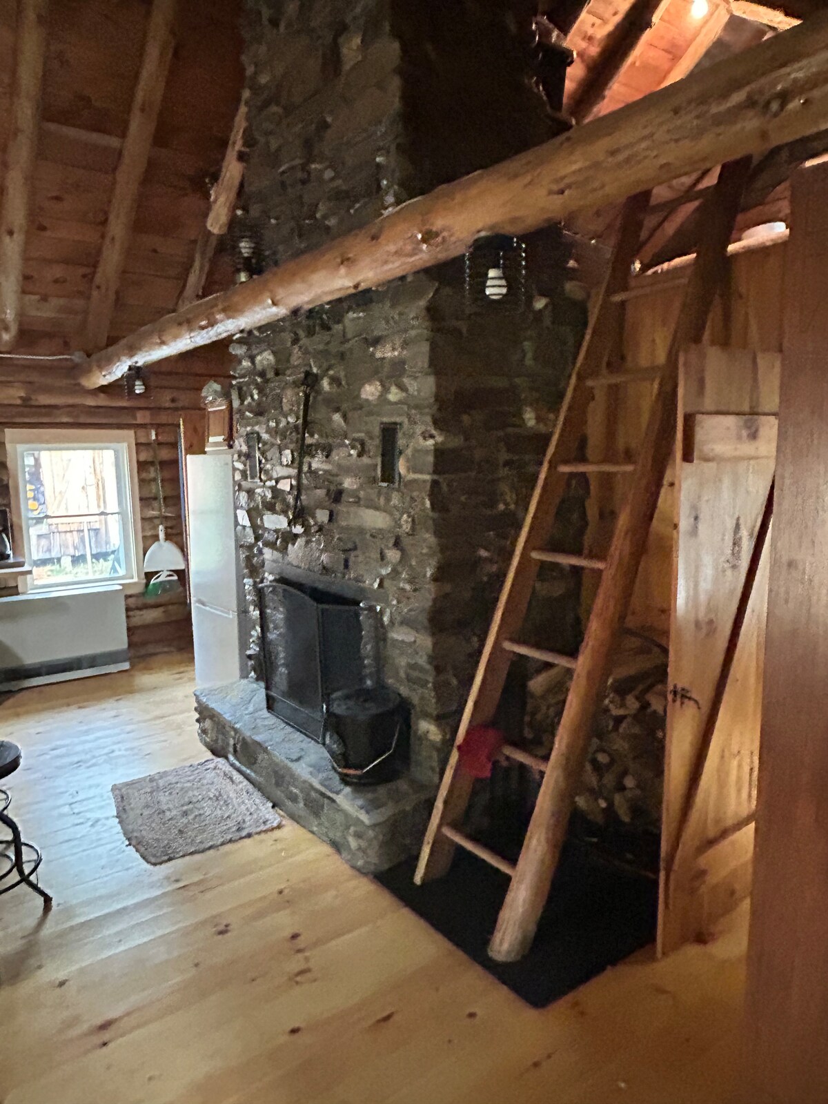 The interior of the log cabin features a rustic stone fireplace, complemented by wooden beams and a natural wood floor. A wooden ladder leads to a loft space, while a cozy seating area and a small decorative rug enhance the cabin's warm aesthetic.