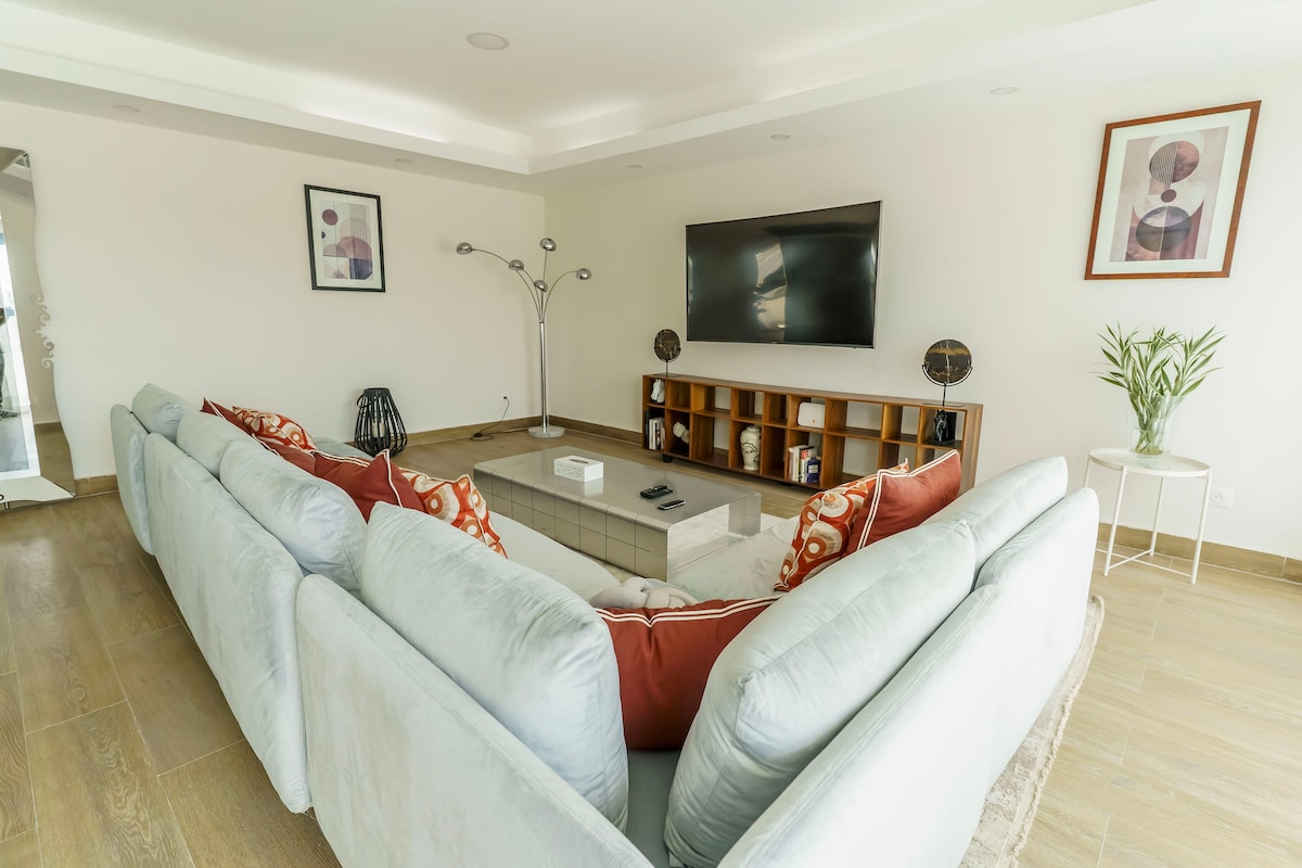 A spacious living area features a large sectional sofa adorned with decorative pillows. A contemporary entertainment unit holds a flat-screen television and decorative items. Natural light fills the room from large windows, while artwork adds a touch of style to the walls.