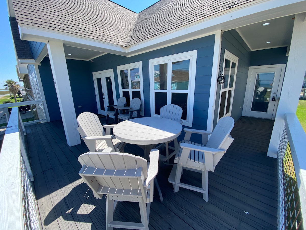 A spacious deck is furnished with a round table surrounded by six adirondack chairs. The blue exterior walls of the house complement the natural surroundings, while large windows provide views of the outdoor area. The deck is illuminated by natural light on a clear day.