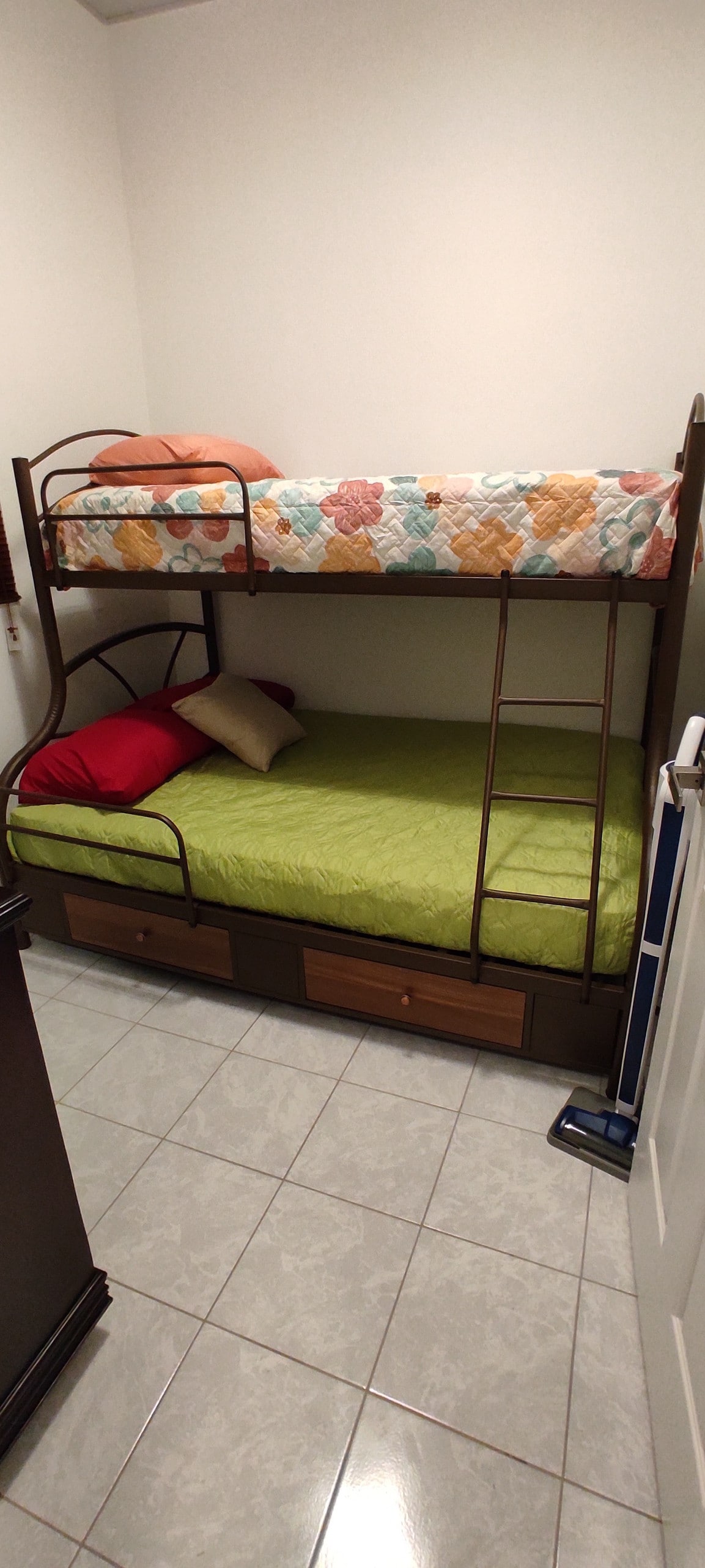 A compact sleeping area features a bunk bed with an upper mattress adorned with a floral-patterned coverlet, and a lower mattress dressed in a vibrant green cover. Two drawers are integrated beneath the bed for storage. The room has light-colored tile flooring.