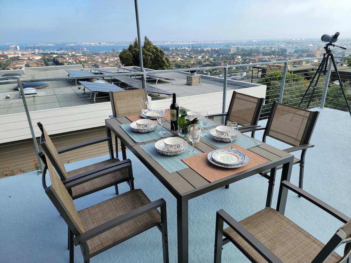 A dining table set for six is positioned on the terrace, overlooking panoramic views of the city and bay. The table is adorned with dinnerware and glasses, complemented by a wine bottle. A telescope is visible in the background, enhancing the scenic experience.