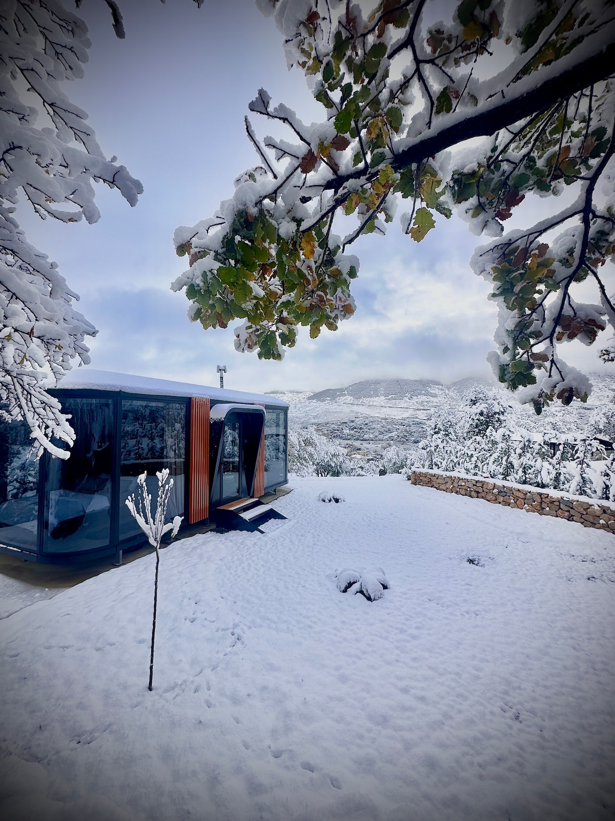 A modern structure is surrounded by a blanket of fresh snow, with snow-covered trees framing the scene. The landscape features rolling hills in the distance, while a clear sky adds a serene ambiance to the wintry setting.