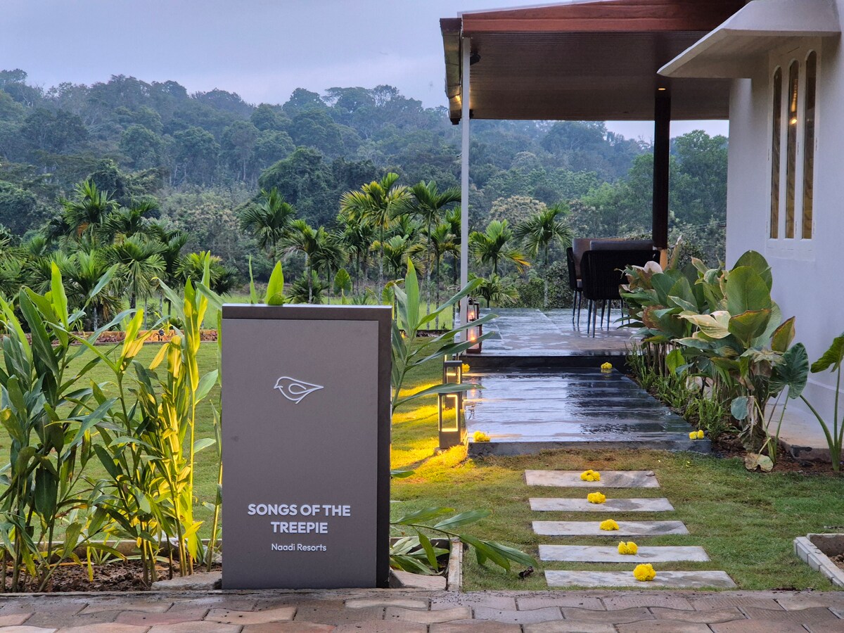 A contemporary entrance is highlighted by a cleanly paved pathway surrounded by lush greenery and vibrant flowers. A sign displaying the name 'SONGS OF THE TREEPIE' stands prominently. The backdrop features tall trees, blending seamlessly with the serene landscape.