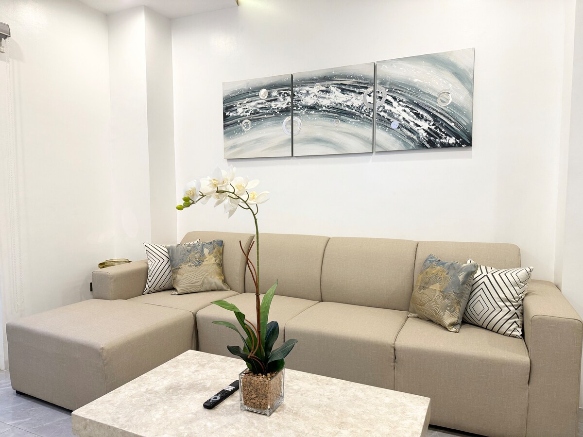 A light and airy living space features a spacious beige sectional sofa adorned with decorative pillows, complemented by a stylish coffee table. A potted orchid adds a touch of greenery, while a three-panel abstract artwork hangs above, enhancing the modern aesthetic.