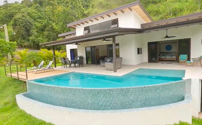 The image showcases Villa Plumeria, featuring an outdoor infinity edge pool extending from the elegant living area. Lounge chairs are positioned by the pool, with a spacious deck overlooking the lush greenery. Large glass doors enhance the connection between indoor and outdoor spaces.