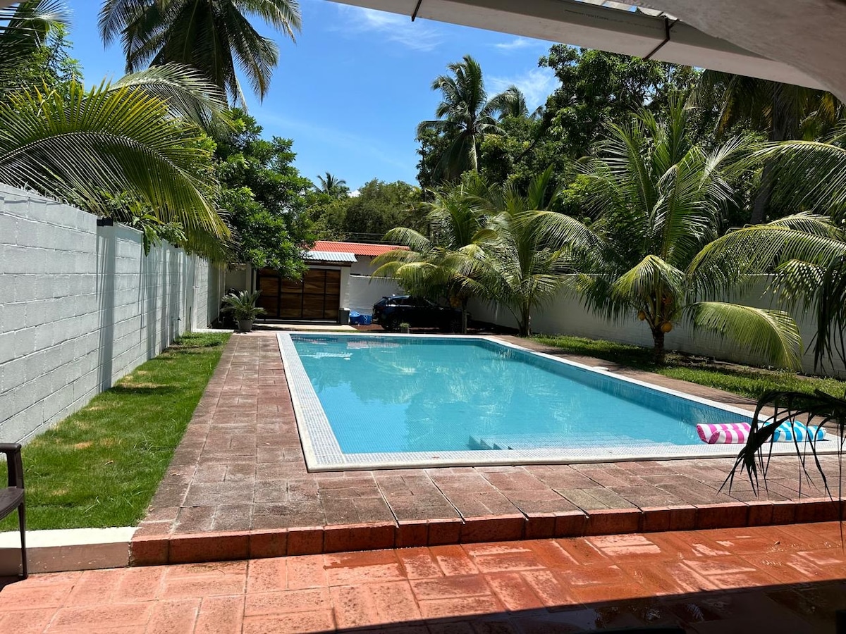 An outdoor area features a large swimming pool surrounded by well-maintained grass and palm trees. A spacious patio edge provides a view of the pool, with sunlight reflecting on the water's surface under a clear blue sky.