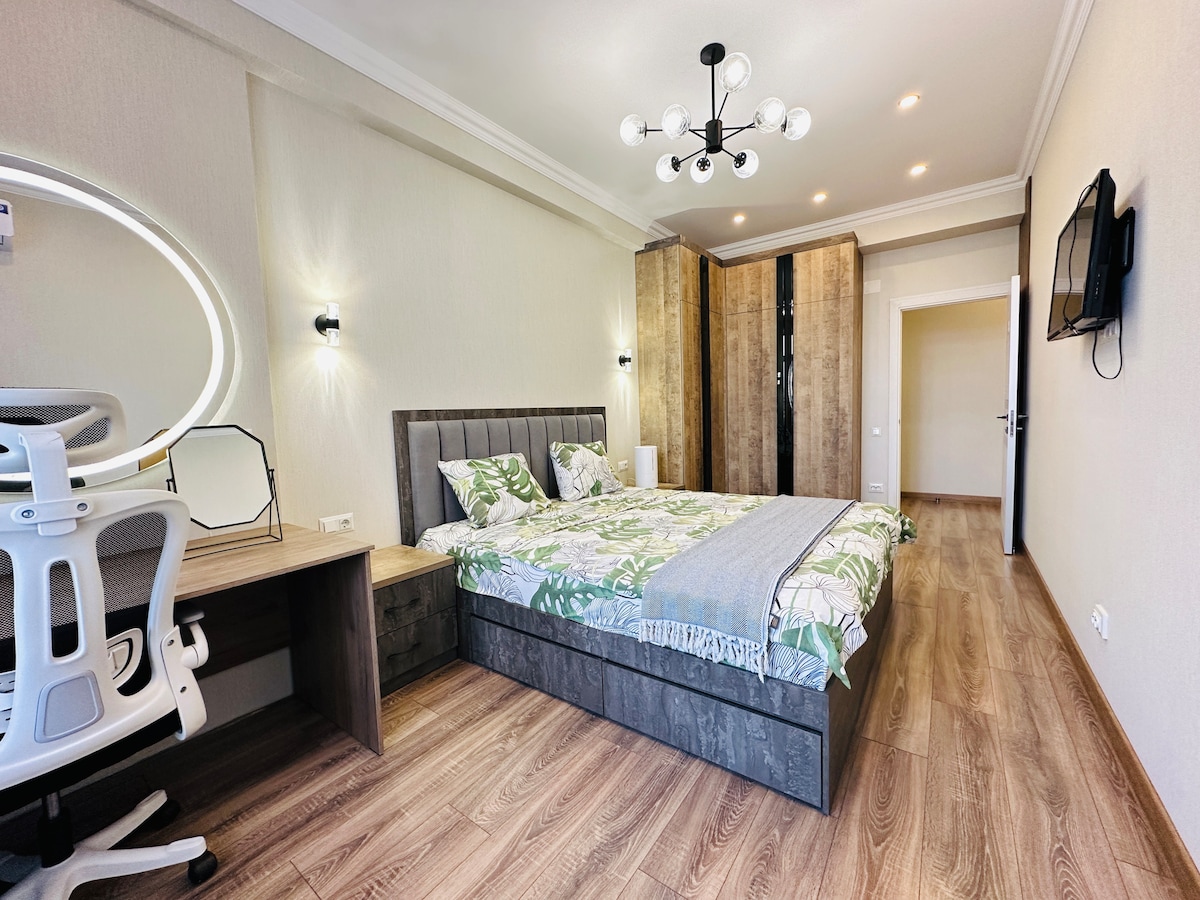 A modern bedroom features a spacious bed dressed in leafy patterned linens. A contemporary desk with a comfortable chair is positioned nearby. Soft ambient lighting is provided by ceiling fixtures and wall sconces, while a stylish wardrobe is visible in the background on a wood-paneled wall.