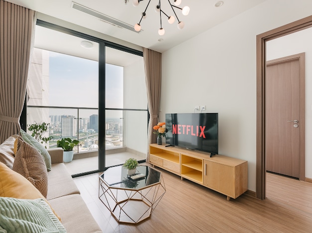 A modern living area features a comfortable sofa complemented by decorative cushions. A television displaying a streaming service is positioned on a wooden media console. Natural light fills the space through large glass doors that lead to a balcony offering city views.