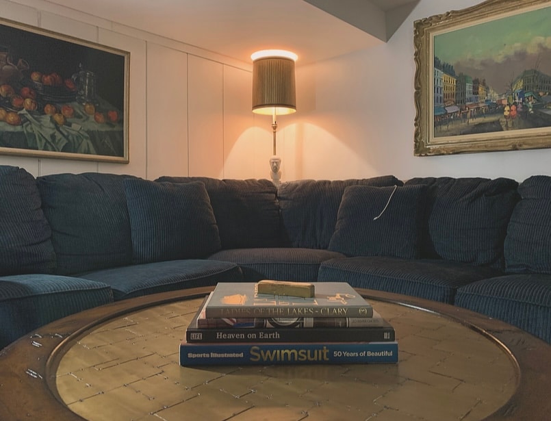 A spacious sectional couch is positioned in a cozy living room, accompanied by a floor lamp providing soft illumination. A coffee table is adorned with several stacked books, while framed artwork enhances the surrounding walls.