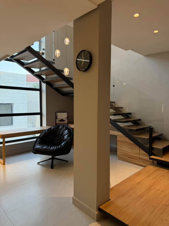 A contemporary indoor space features a modern staircase with glass railings, leading to upper levels. A sleek black chair is positioned next to a wooden table, creating a cozy nook. Soft lighting is provided by pendant lights hanging overhead, while a wall clock adds functionality.