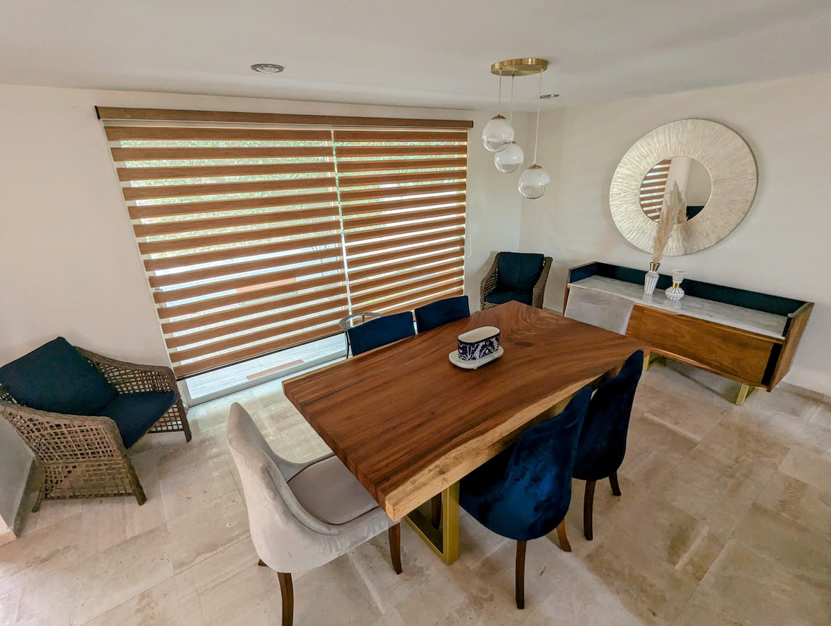 A dining area is presented with a large wooden table surrounded by six chairs in various upholstery. Natural light filters through horizontal blinds, illuminating the space. A decorative mirror is mounted on the wall, and a sideboard adds functionality to the room.