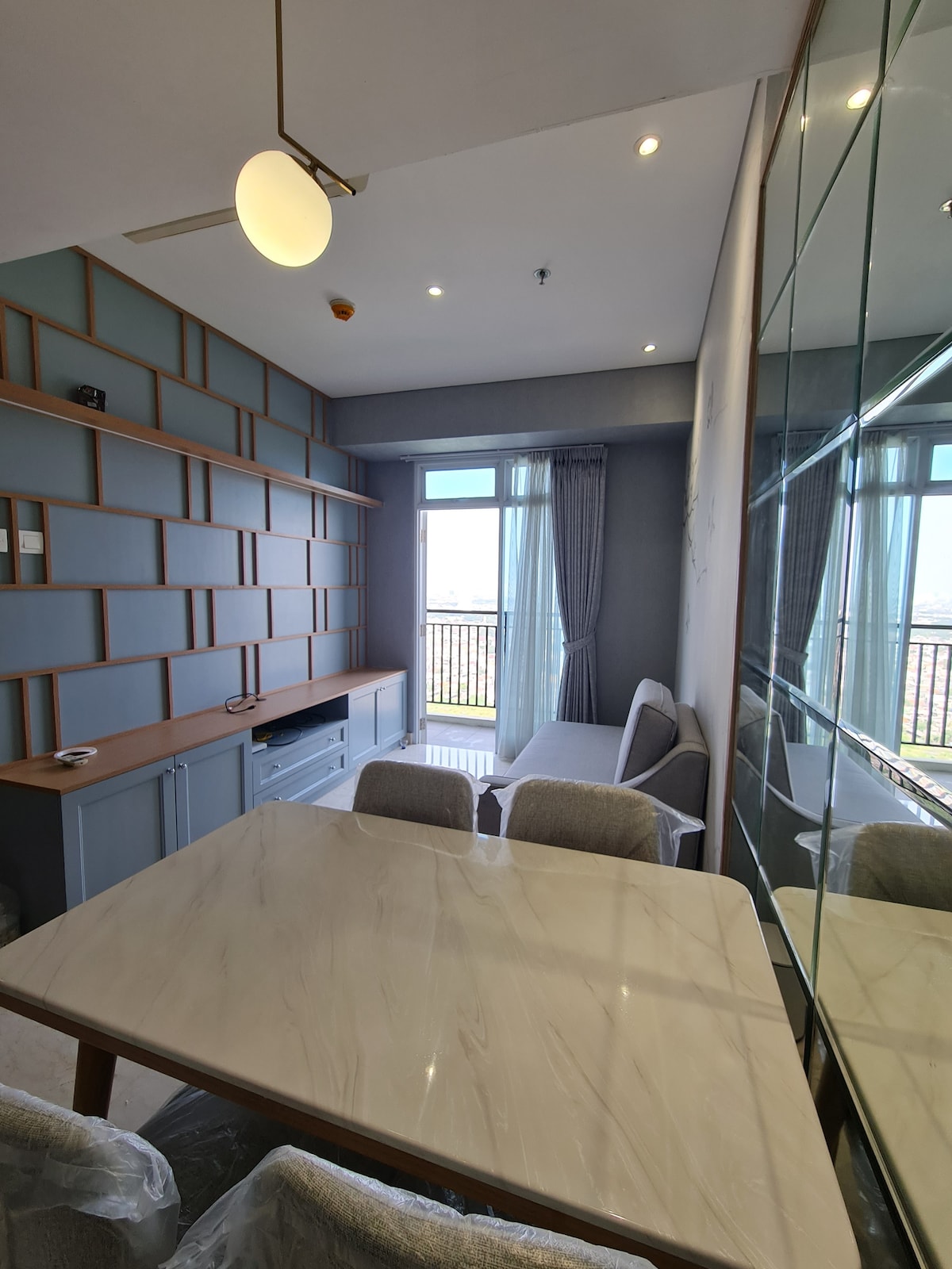 A modern living area features a light wood dining table surrounded by four upholstered chairs. A sleek entertainment unit and a large mirror enhance the space, while natural light enters through the tall window dressed with sheer curtains.