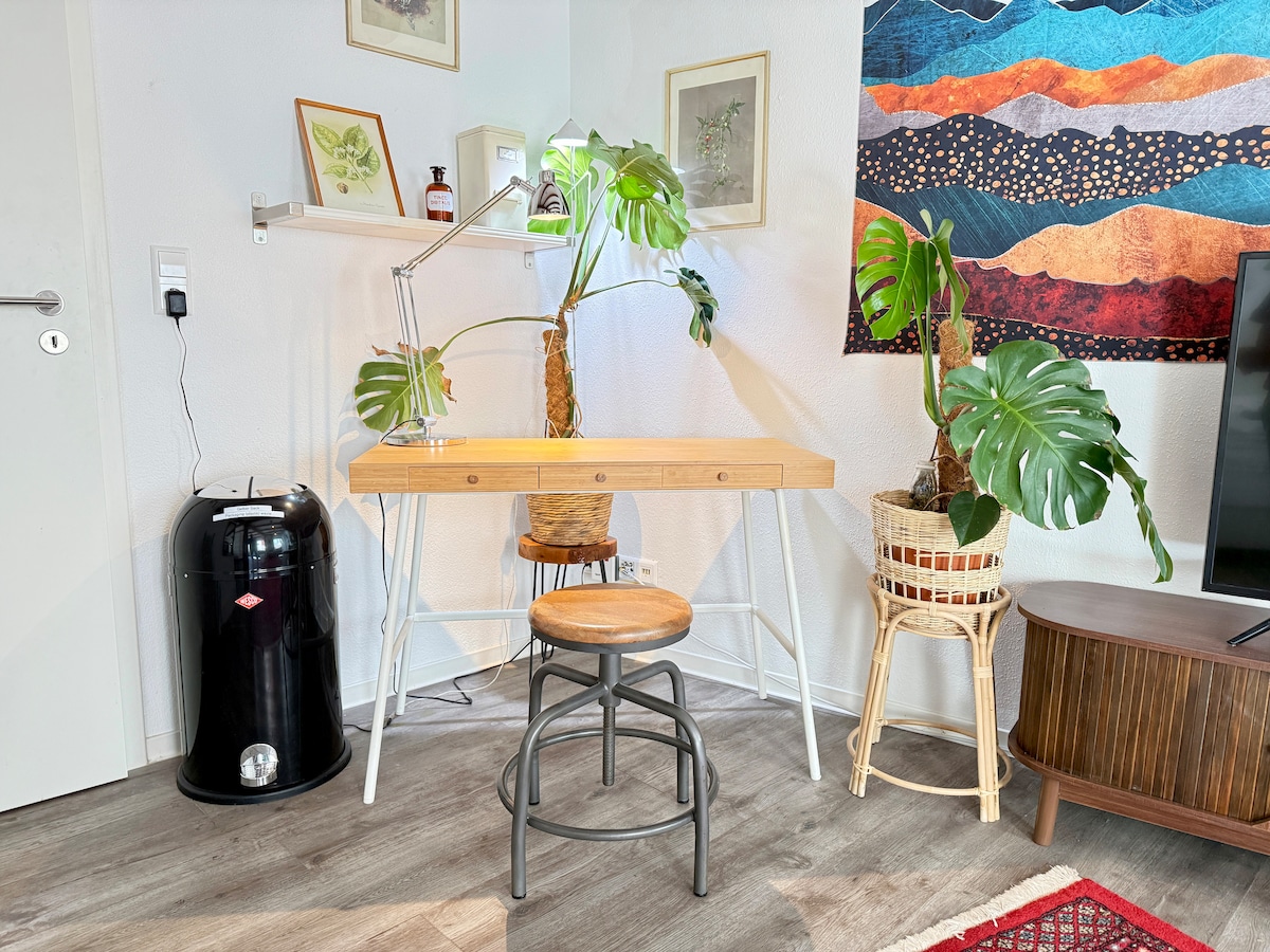 A small workspace is featured with a wooden desk and a metal stool. Two potted plants add a touch of greenery. A black trash bin stands nearby, while a colorful wall hanging and a television on a side table complete the inviting setting.