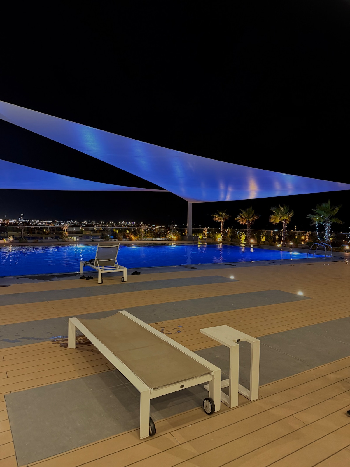A tranquil pool area is highlighted with soft lighting at night, featuring crystal-clear water. Comfortable lounge chairs and a small table are positioned on a wooden deck, with modern umbrellas providing shade. Palm trees dot the surrounding area, enhancing the serene ambiance.
