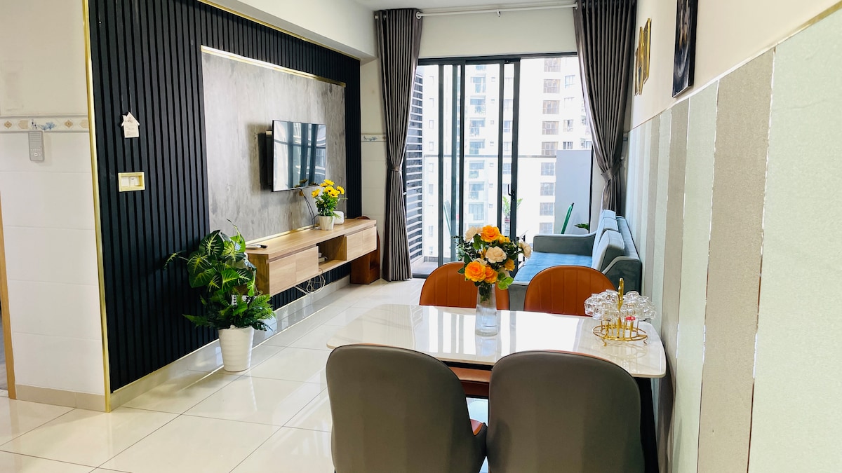 A modern living space features a comfortable seating area with a blue sofa facing a wall-mounted television. A dining table is set with elegant tableware and a vase of fresh flowers. Floor-to-ceiling windows provide natural light, enhancing the open feel of the room.