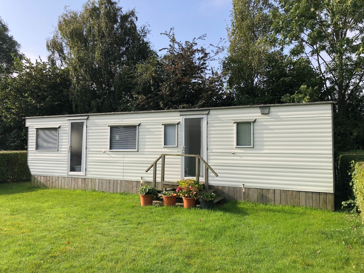 A well-maintained mobile home is surrounded by lush green grass, featuring multiple windows that provide natural light. A small wooden staircase leads to the entrance, where potted plants add a touch of color to the exterior.