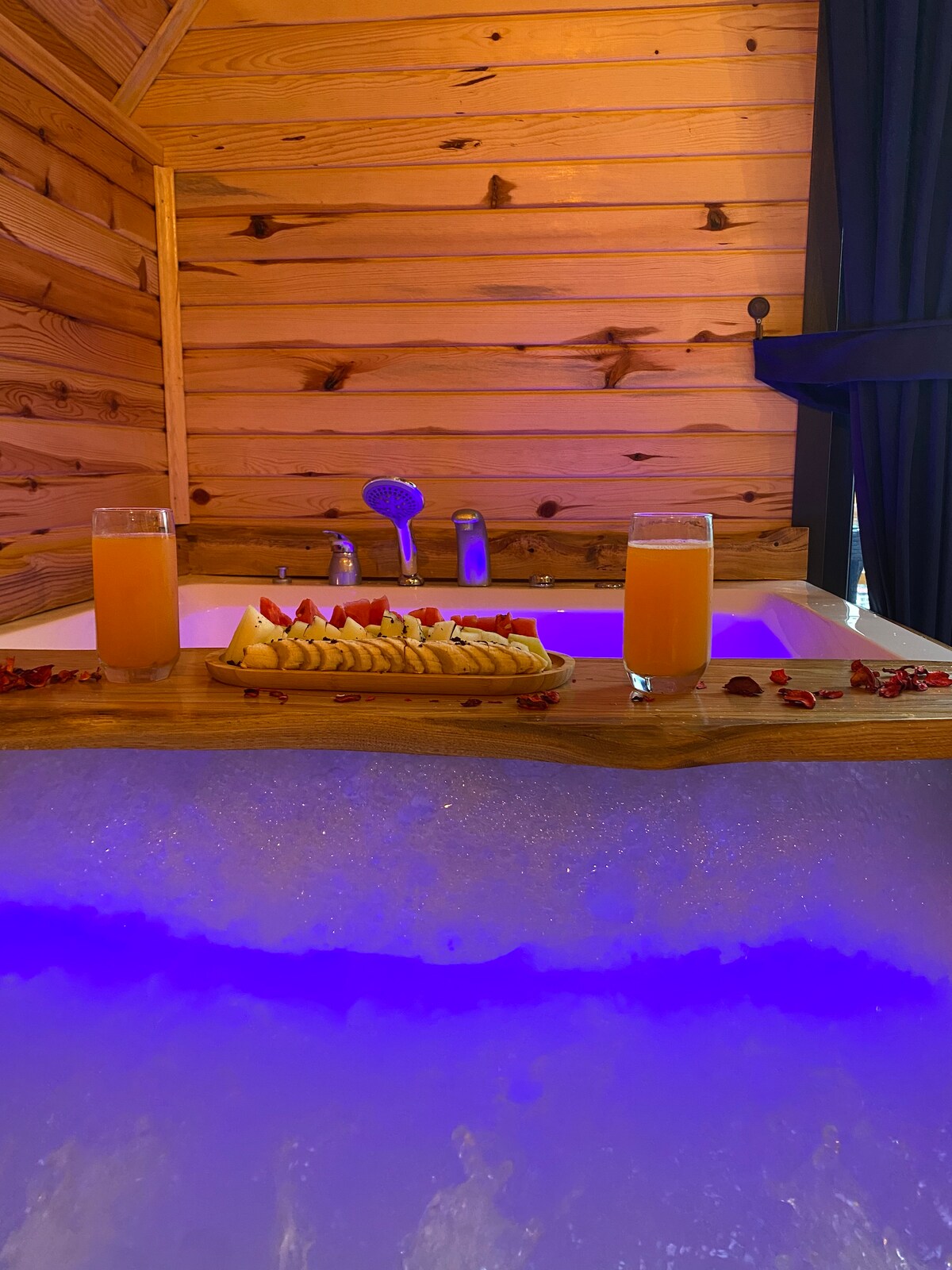 A relaxing spa area is highlighted by a large tub surrounded by wooden walls. Brightly colored beverages are placed on the edge, alongside a tray of fresh fruit. Soft lighting enhances the soothing atmosphere, suggesting a tranquil retreat.