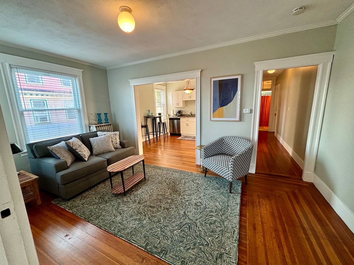 The living area features a comfortable sofa accompanied by decorative pillows. A wooden coffee table occupies the center space, complementing a patterned area rug. Light from a nearby window brightens the room, which connects to the kitchen and hallway.