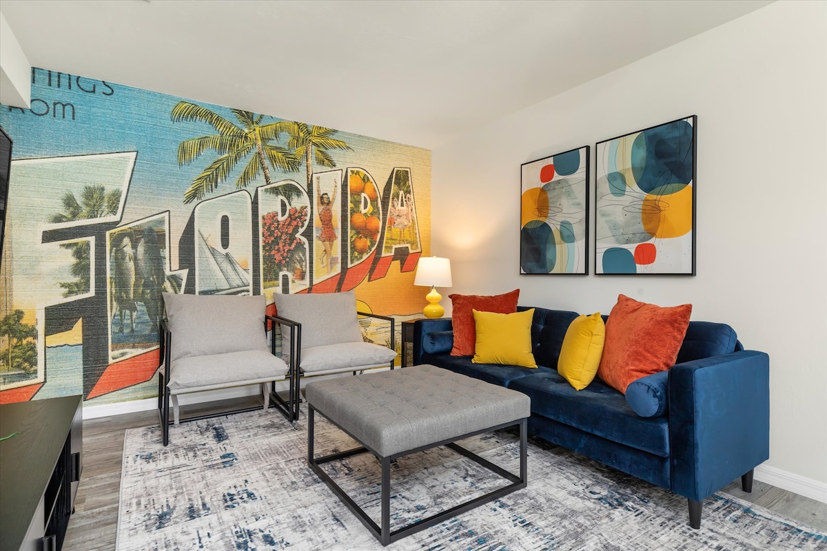 A vibrant living area features a blue sofa adorned with colorful cushions, offering a welcoming space to relax. Two gray armchairs are positioned beside a modern coffee table. A large mural depicting a tropical scene enhances the ambiance, complemented by contemporary artwork on the adjacent wall.
