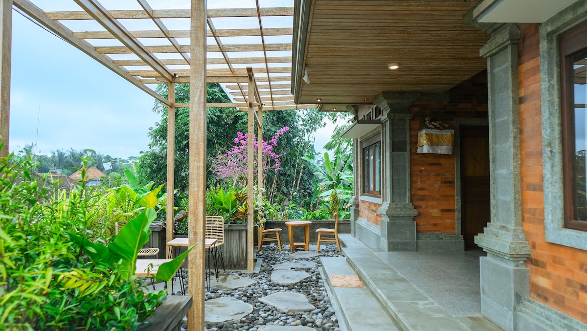 An outdoor terrace features a wooden pergola offering partial shade, complemented by lush greenery and vibrant pink flowers. Comfortable seating arrangements are visible, creating an inviting space to relax and enjoy the surroundings. A stone pathway leads through the garden area, enhancing the natural ambiance.