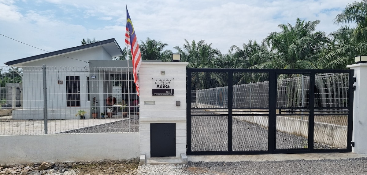 A modern house is seen behind a gated entrance, featuring a sleek black metal gate and a flag displayed prominently. The front yard is gravelled, surrounded by low fencing and tropical greenery, enhancing the peaceful environment.