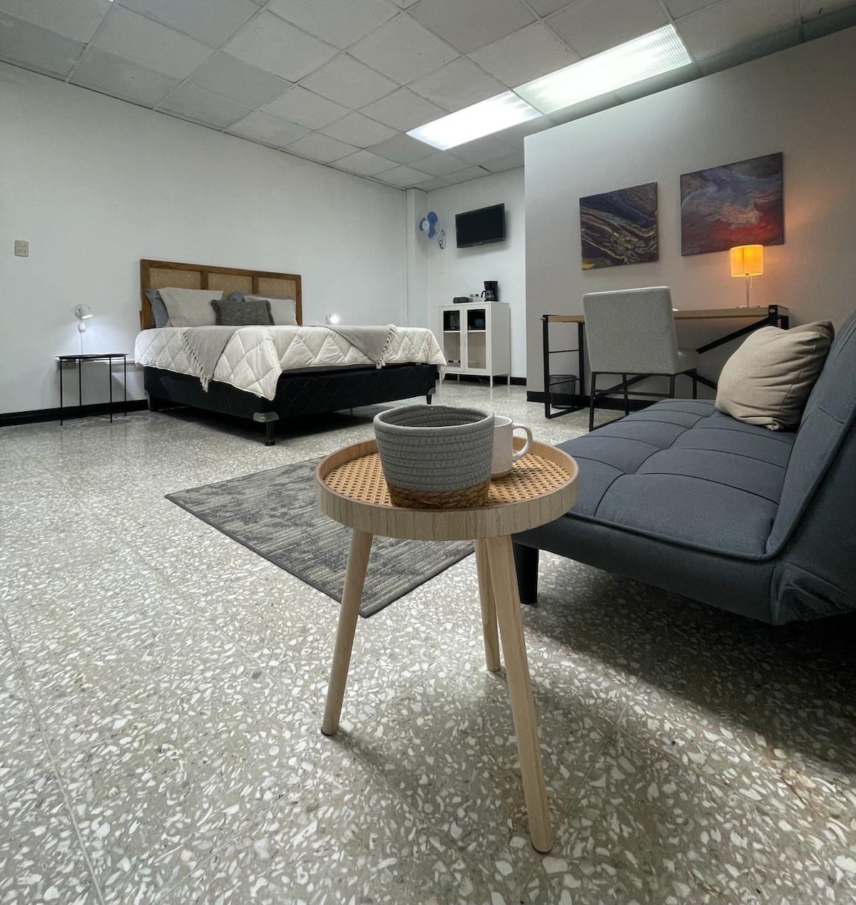A spacious studio features a large bed with a quilted cover and bedside lamps on either side. A small table is positioned in the foreground with a cup and a cozy sofa is visible nearby. A kitchenette and a wall-mounted TV are also present.