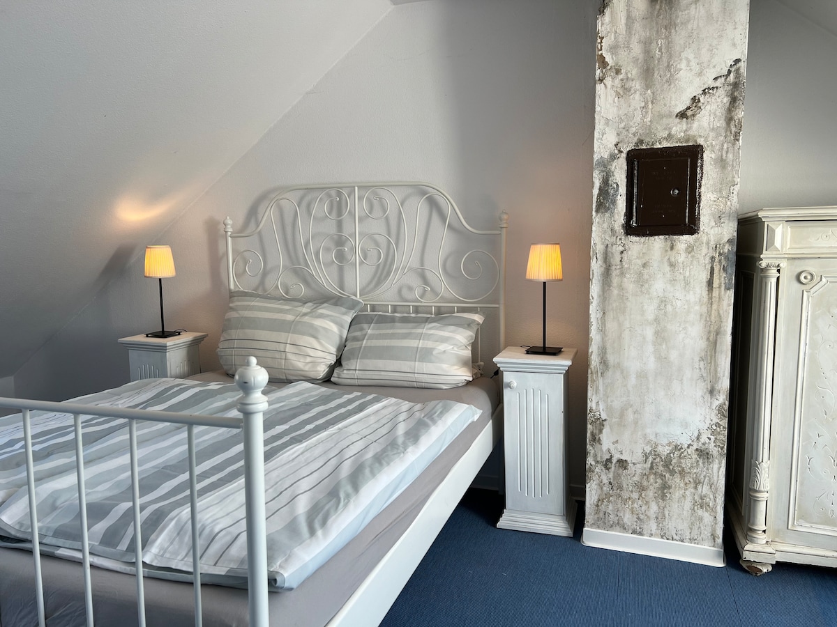 A well-appointed bedroom features a white metal bed with soft linens. Two bedside lamps provide gentle lighting, and a vintage cabinet is positioned in the corner. The walls are painted in a light tone, complemented by a textured concrete feature, enhancing the space's character.