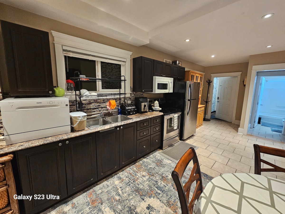 A well-equipped kitchen features dark cabinetry and a marble countertop, providing ample workspace. Modern appliances, including a microwave and refrigerator, are present. A dining table is positioned nearby, and a doorway leads to a brightly lit area, enhancing the overall functionality of the space.