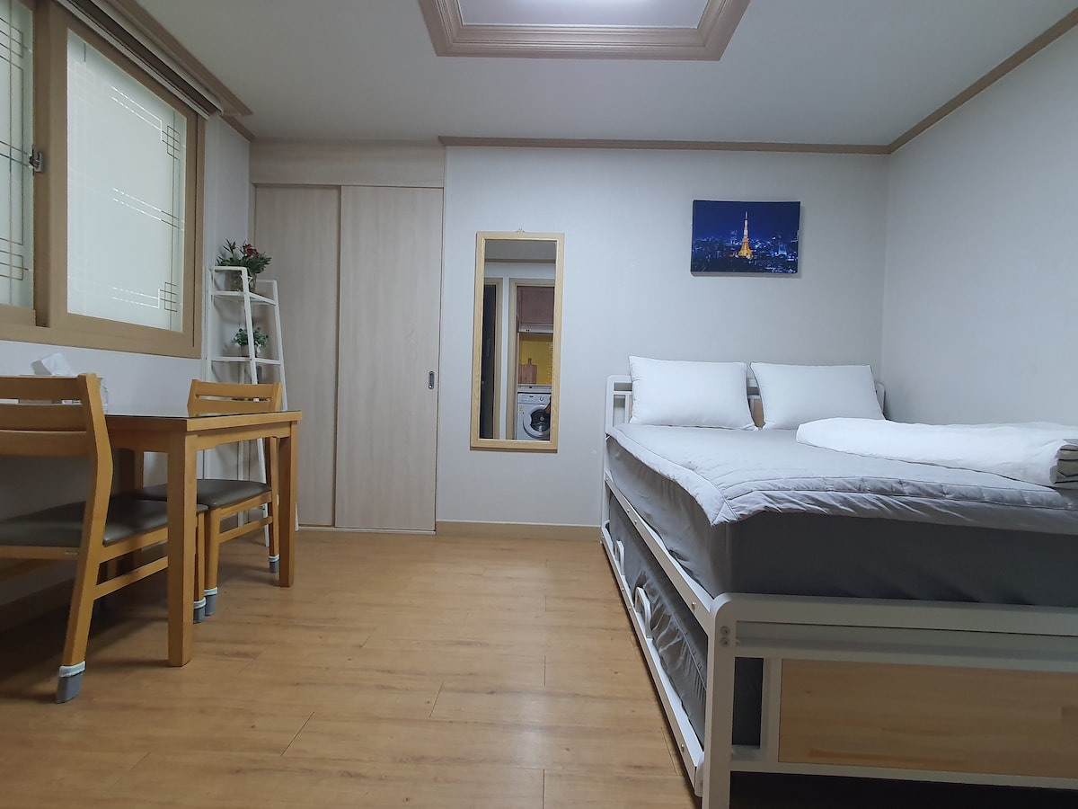 A neatly arranged room features two queen beds with crisp white linens. A small dining table with two chairs occupies one corner. Natural light enters through a window, and a mirror reflects the space. The warm wooden floor complements the light-colored walls.