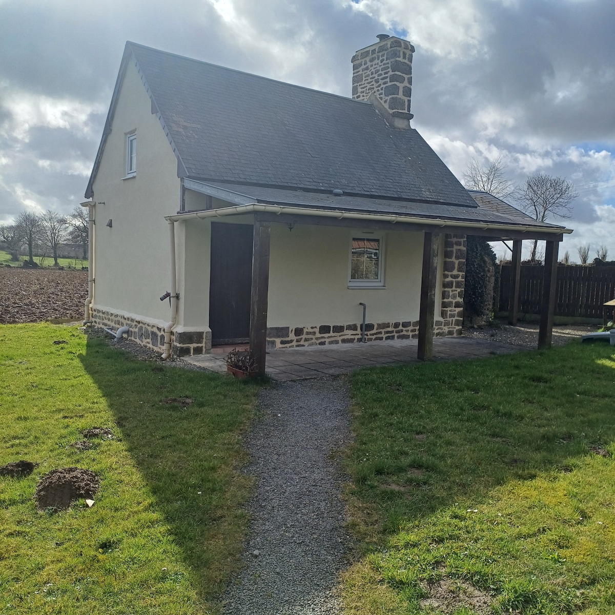 A charming cottage is presented with a light-colored facade and a prominent stone chimney. The pathway is surrounded by a grassy area, leading to the entrance. Fields are visible in the background under a partly cloudy sky.