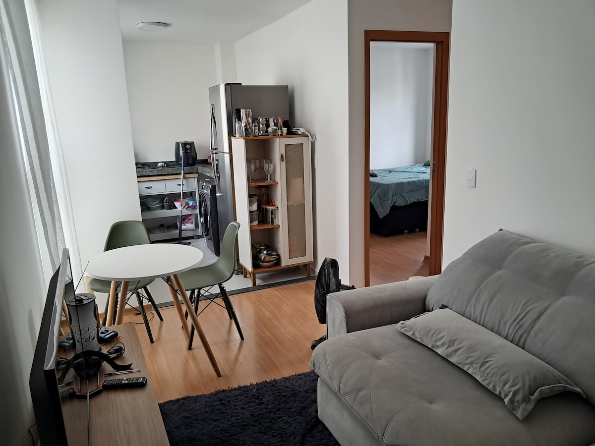 A modern living area features a comfortable grey sofa and a round dining table surrounded by green chairs. A flat-screen television is positioned on a low stand. The kitchen area, visible in the background, includes a sleek appliance setup. Light pours in through the window.