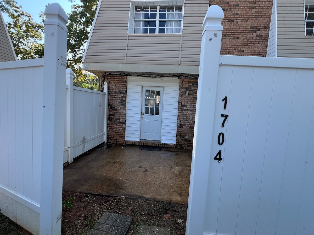 A private entrance is framed by white fencing, leading to a door with glass panels. The ground is paved with concrete, and the address number is clearly displayed on the fence. The area appears clean and well-kept.
