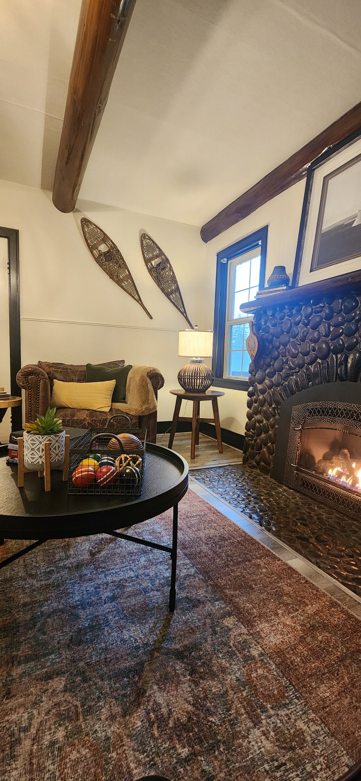 A cozy living area is highlighted by a rock gas fireplace, surrounded by rich wooden beams. A leather couch offers seating, complemented by a round table adorned with decorative items. Natural light fills the space through a window, creating a warm ambiance.