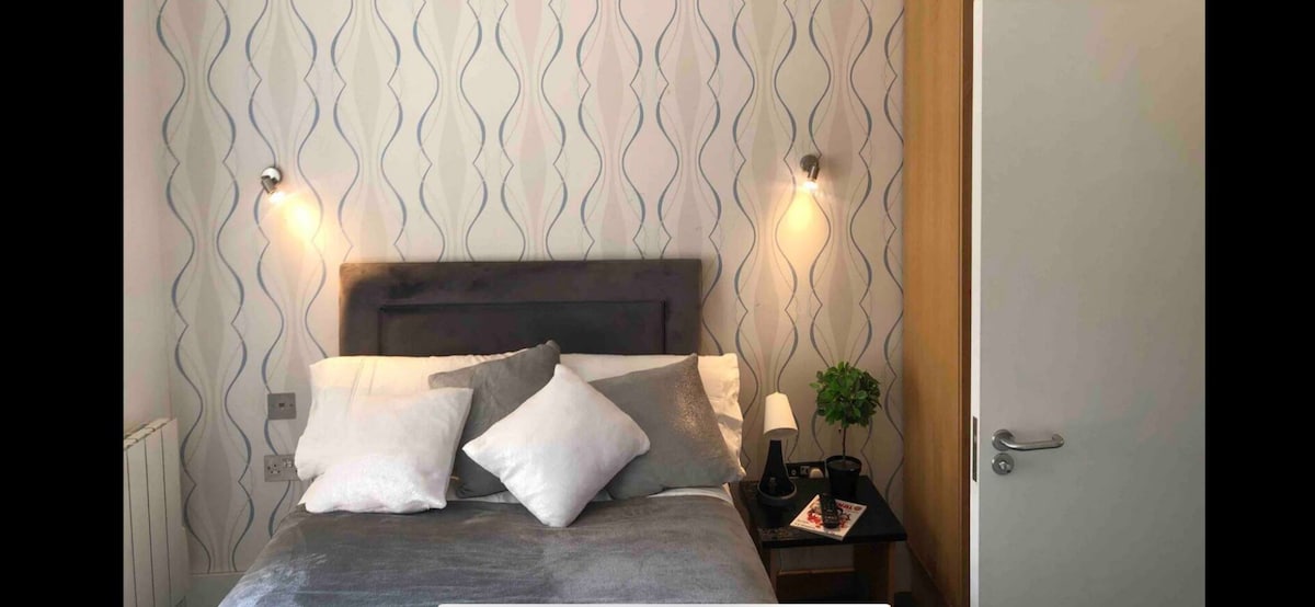 A cozy sleeping area is presented with a plush bed, featuring soft grey linens and an assortment of decorative pillows. A small bedside table holds a lamp and a potted plant. The wall displays a subtle, patterned design, complementing the warm lighting from sconces.