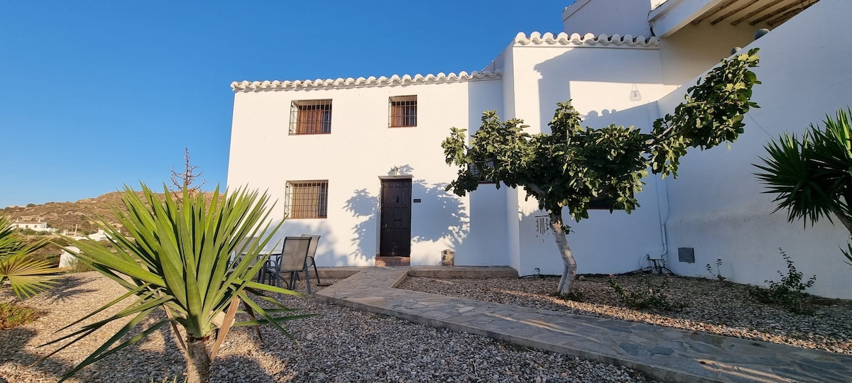 The exterior of a white, two-story house is presented, featuring a simple door and several windows. A stone pathway leads to the entrance, bordered by gravel and greenery. Sunlight highlights the clear blue sky and the surrounding mountainous landscape.