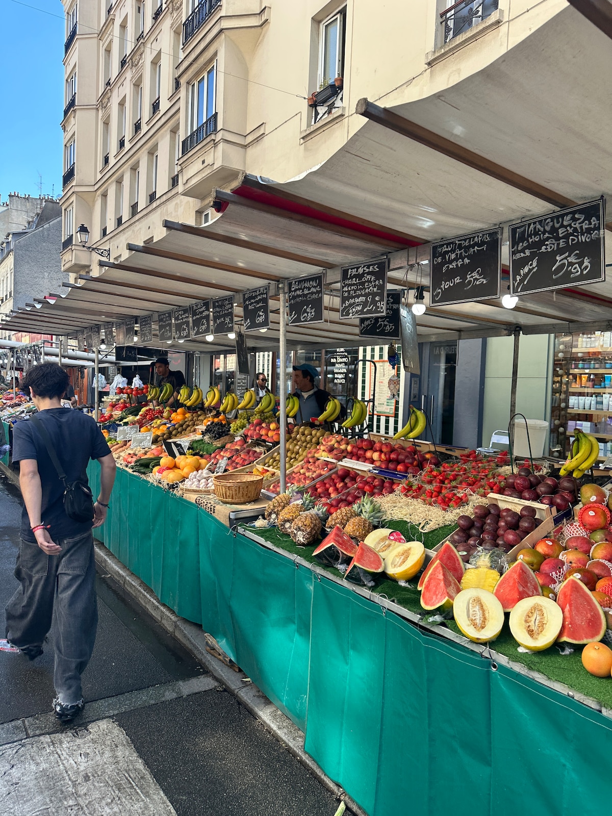 A bustling market scene features rows of colorful stalls displaying a variety of fresh fruits, including watermelons, bananas, and grapes. The green tablecloths provide a vibrant contrast against the assortment of produce, while handwritten signs detail the offerings. A passerby walks along the market path.