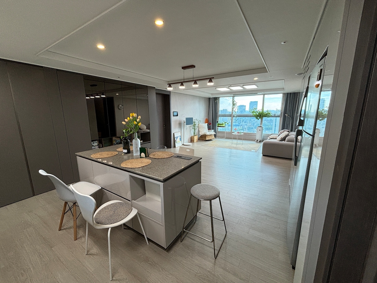 An open-plan living area showcases a modern kitchen island with seating. Natural light fills the space, highlighting sleek cabinetry and a comfortable lounge area. Floor-to-ceiling windows frame scenic views, blending indoor and outdoor environments. Decorative plants add a touch of freshness.
