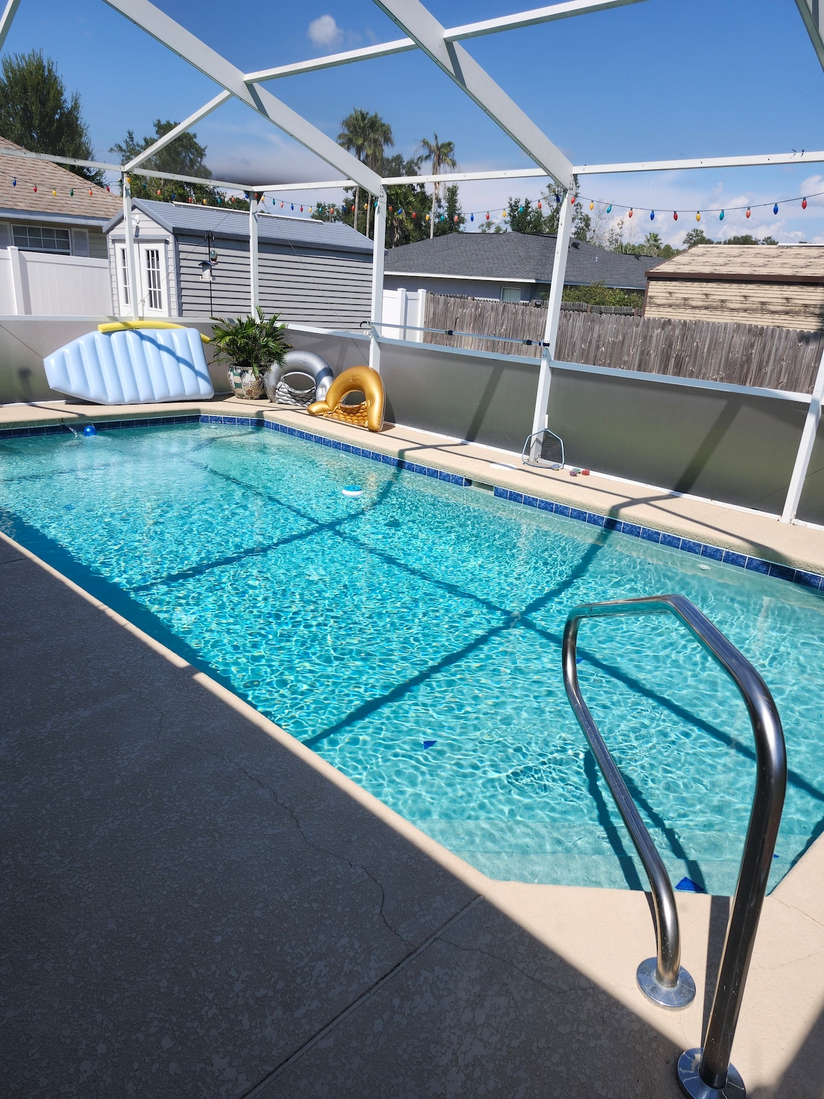 A rectangular swimming pool is visible, surrounded by a patio area. Sunlight reflects off the water surface, creating vibrant patterns. A safety railing leads into the pool. Loungewear inflatables are placed on the pool's edge, adding a fun touch to the outdoor space.