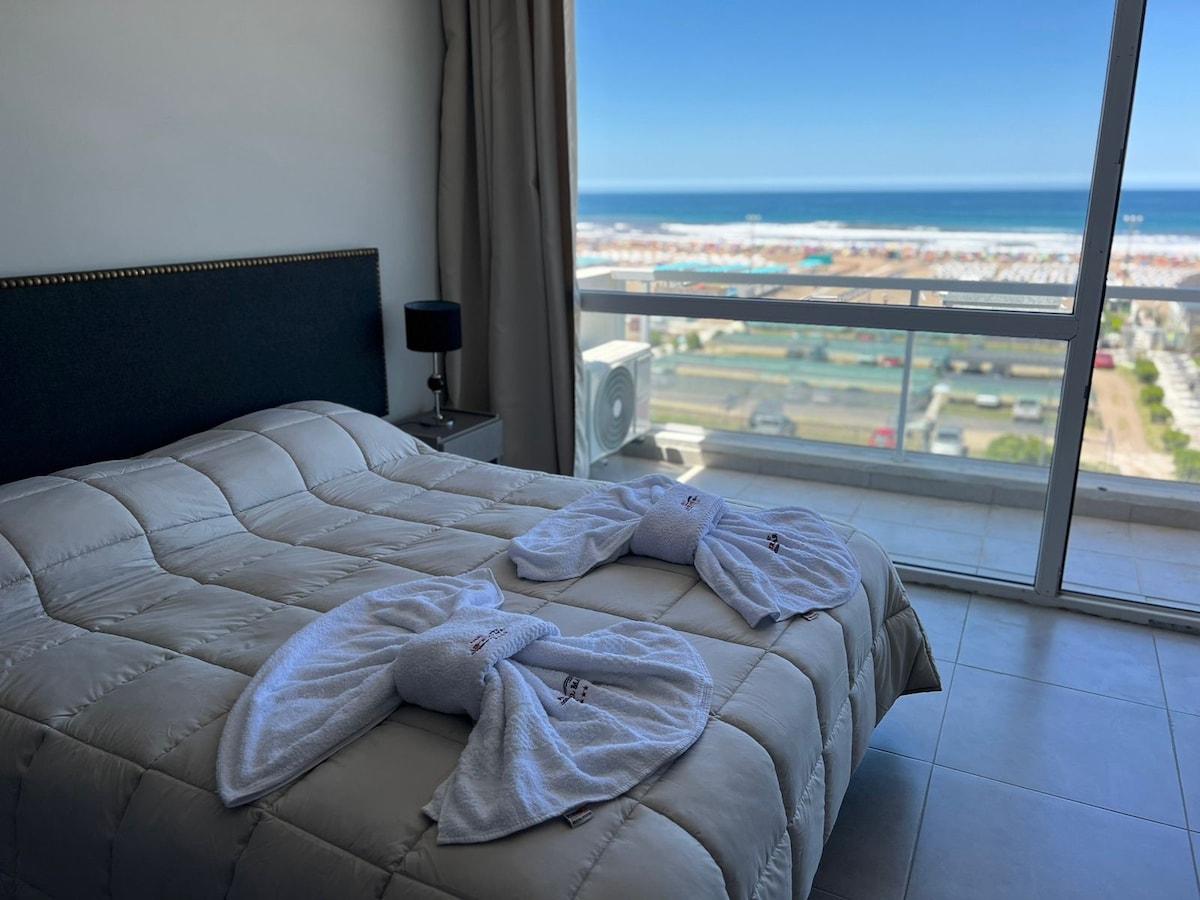 A spacious sleeping area features a king-sized bed covered with a light quilt, complemented by decorative towels styled as swans. Large windows open to a stunning ocean view, allowing natural light to brighten the room, with a cozy bedside lamp providing additional illumination.