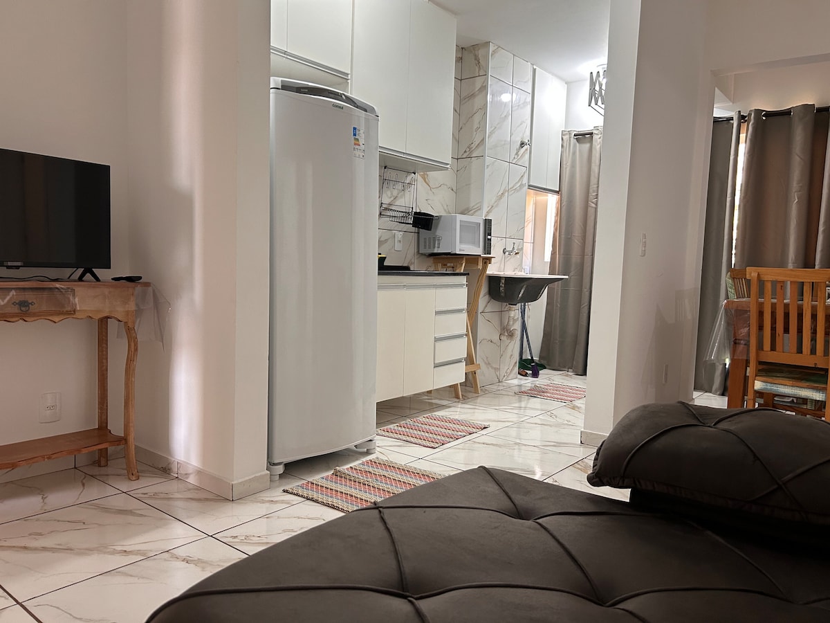 A modern living space is shown, featuring a comfortable bed with a quilted dark cover. A kitchenette with white cabinetry and a shiny refrigerator is visible. A dining area is positioned in the background, with a wooden table and chairs, while a TV stands on a side table.