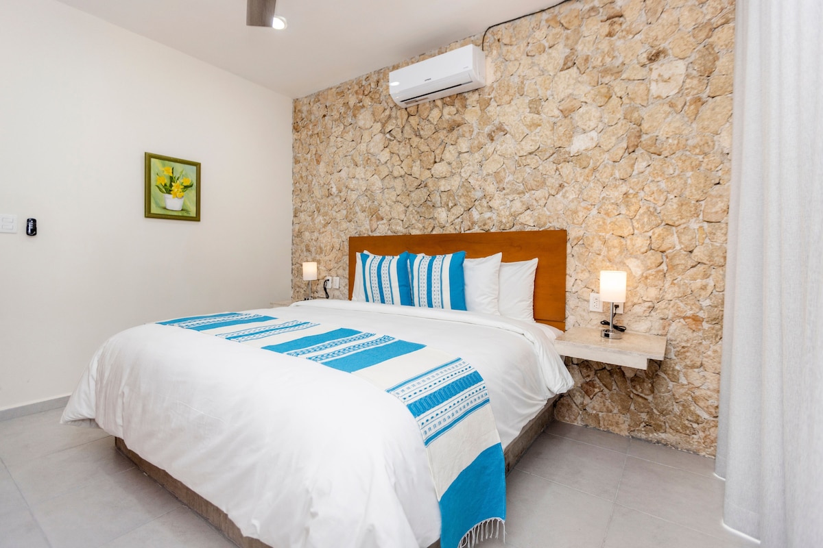 A spacious bedroom features a neatly made bed with a blue and white decorative throw. The stone wall adds texture, while soft lighting from bedside lamps creates a calm ambiance. An air conditioning unit is mounted above, ensuring comfort in the room.
