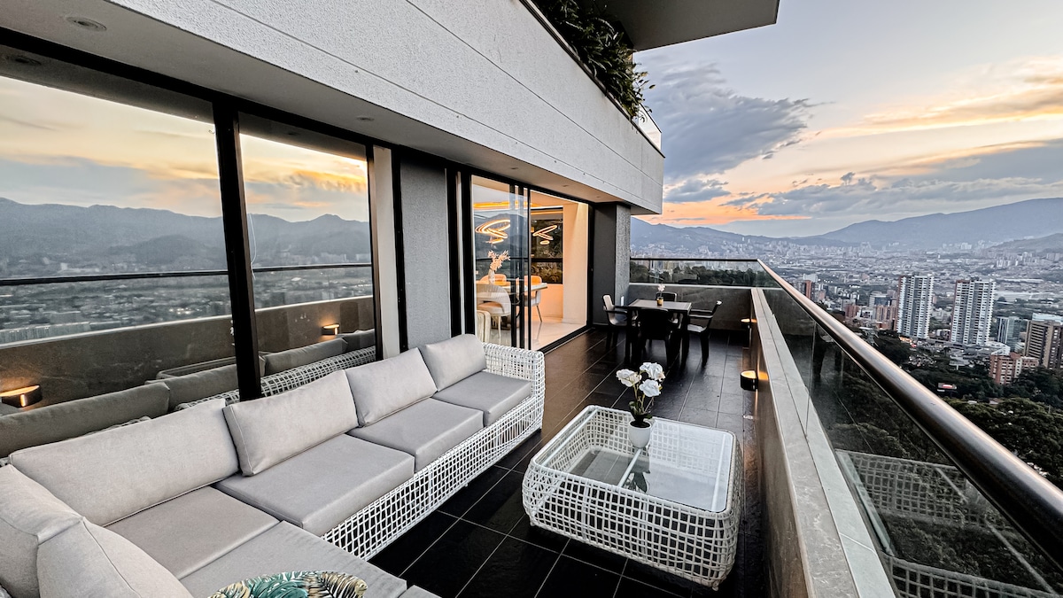 A spacious balcony features modern wicker seating and a glass-topped coffee table, facing sweeping views of the city and mountains. The scene is enhanced by warm evening light, creating an inviting space for relaxation or social gatherings.