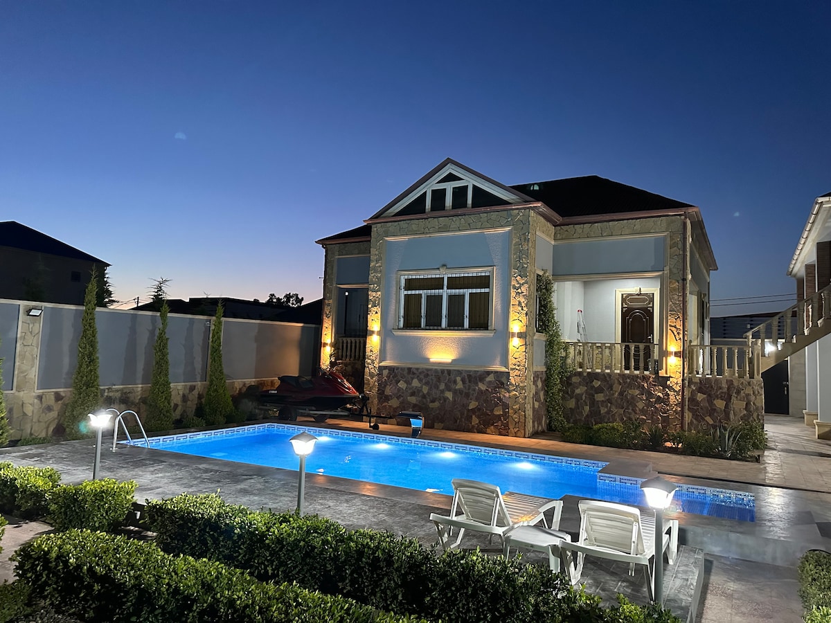 A spacious villa is seen with a clear blue pool illuminated at night. Soft lighting highlights the exterior stonework. Sun loungers are positioned around the pool, and well-maintained greenery adds a touch of nature to the setting. The sky transitions from deep blue to twilight.