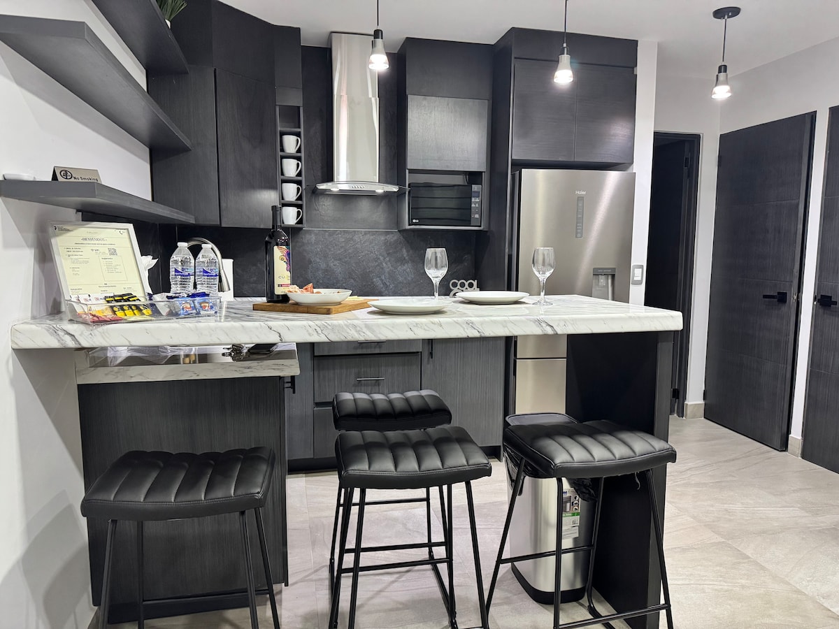 A modern kitchen features dark cabinetry and sleek appliances, including a microwave and refrigerator. A marble countertop serves as a breakfast bar with four black stools positioned underneath. The space is illuminated by pendant lights, creating a bright and functional area.