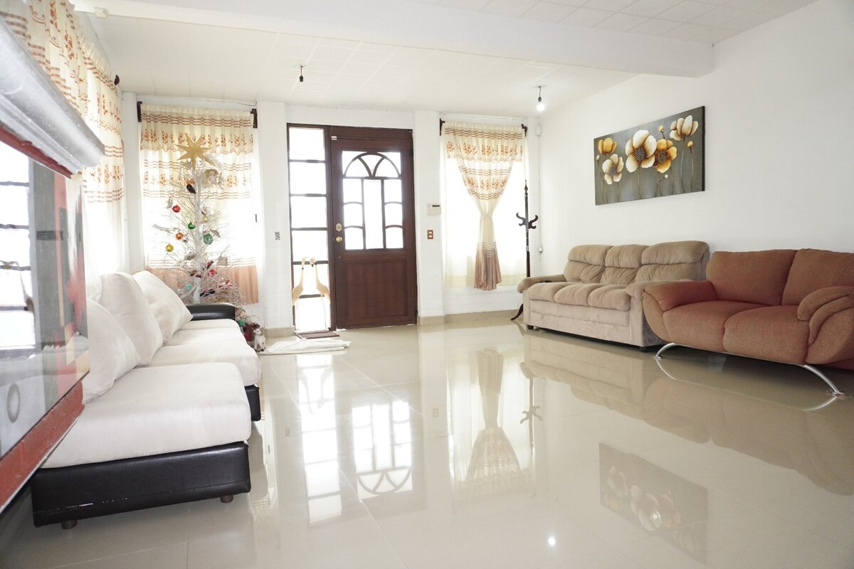 A spacious living area features a combination of comfortable seating options, including a white sofa and a brown sectional. Natural light enters through large windows dressed with light curtains, while a decorative wall art piece adds a touch of elegance. The tiled floor reflects the room's bright ambiance.